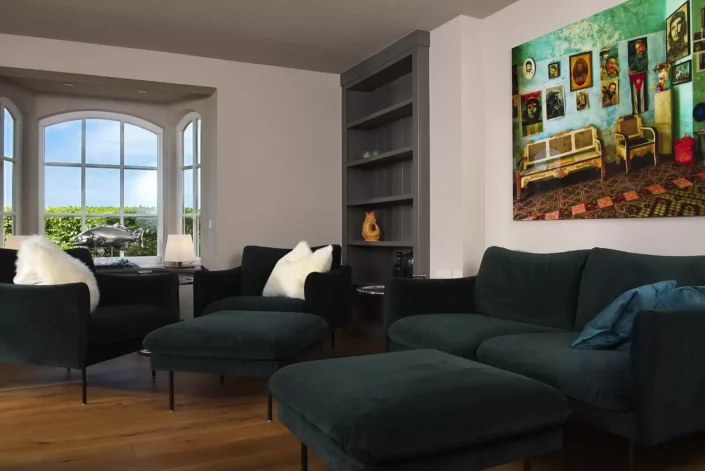 Modernes Wohnzimmer einer Sylter Immobilie mit dunkelgrünen Samtsofas, einem großen Wandbild und einem hellen Erkerfenster mit Blick ins Grüne.