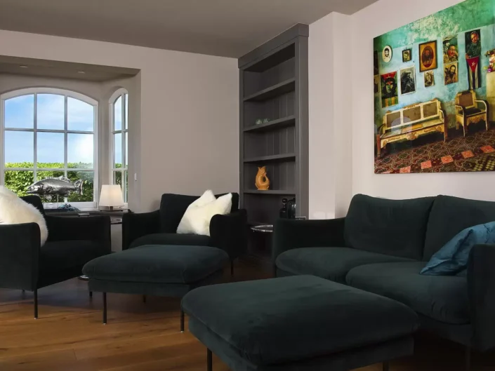 Modernes Wohnzimmer einer Sylter Immobilie mit dunkelgrünen Samtsofas, einem großen Wandbild und einem hellen Erkerfenster mit Blick ins Grüne.