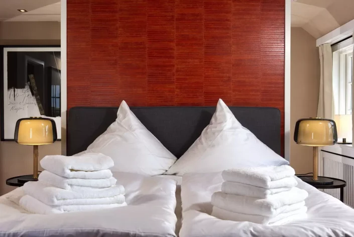 Ein stilvolles Schlafzimmer in einer Sylter Immobilie mit roter Akzentwand, zwei Nachttischlampen und frisch gefalteten weißen Handtüchern auf dem Bett.