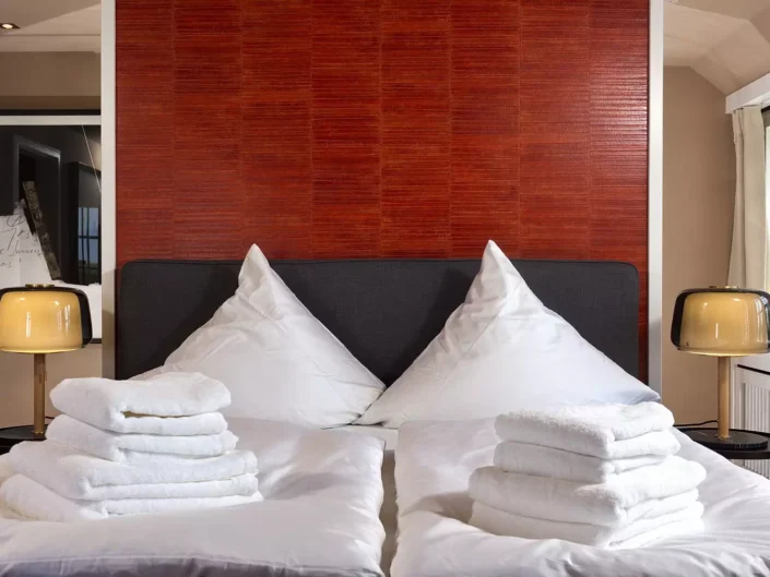 Ein stilvolles Schlafzimmer in einer Sylter Immobilie mit roter Akzentwand, zwei Nachttischlampen und frisch gefalteten weißen Handtüchern auf dem Bett.