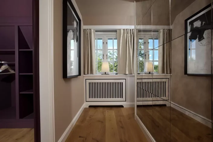Eleganter Ankleidebereich in einer Sylter Immobilie mit Holzboden, Einbauschränken, großem Spiegel und einem hellen Fenster mit Vorhängen.