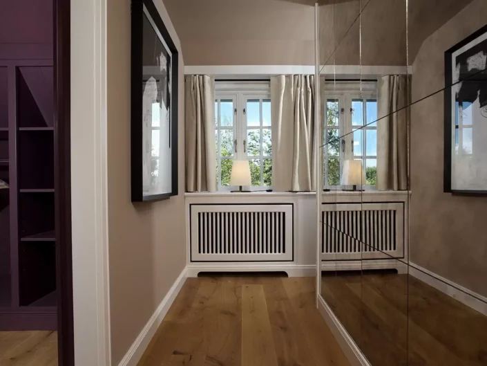 Eleganter Ankleidebereich in einer Sylter Immobilie mit Holzboden, Einbauschränken, großem Spiegel und einem hellen Fenster mit Vorhängen.