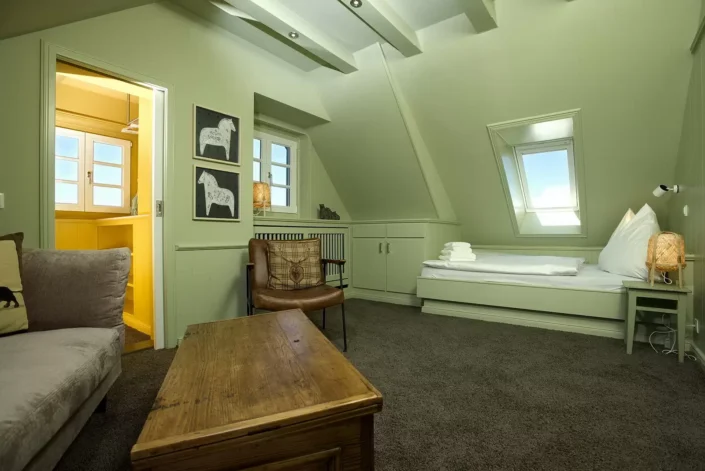 Ein gemütliches Schlafzimmer in einer Immobilie auf Sylt mit grünen Wänden, Dachschrägen, einem Bett, einem Ledersessel und einem Holztisch.