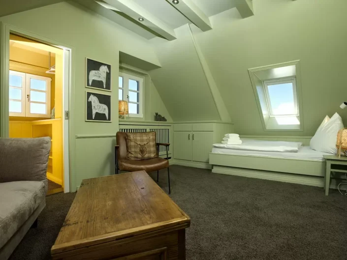 Ein gemütliches Schlafzimmer in einer Immobilie auf Sylt mit grünen Wänden, Dachschrägen, einem Bett, einem Ledersessel und einem Holztisch.