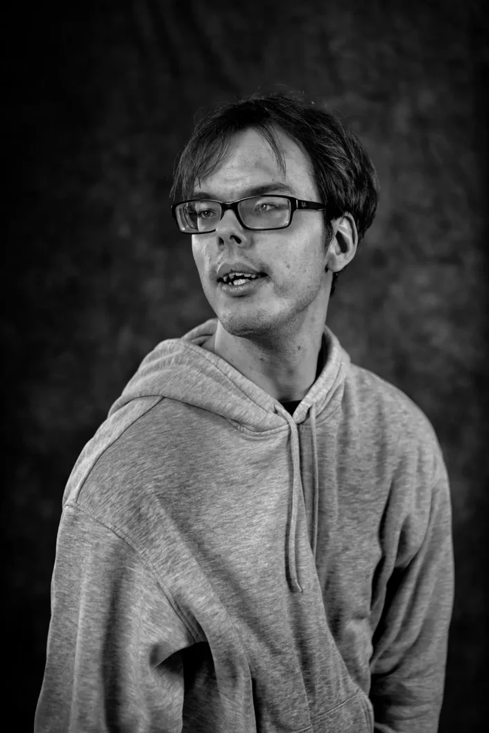 Schwarz-Weiß-Porträt eines jungen Mannes mit Brille und grauem Kapuzenpullover, der auf Sylt vor einem dunklen Hintergrund fotografiert wurde.