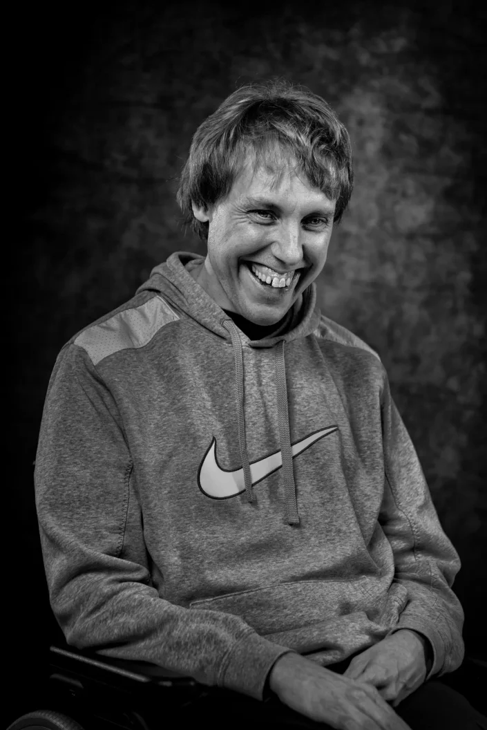 Schwarzweiß Porträt eines lächelnden Mannes in einem grauen Nike Kapuzenpullover vor einem dunklen, strukturierten Hintergrund auf Sylt.