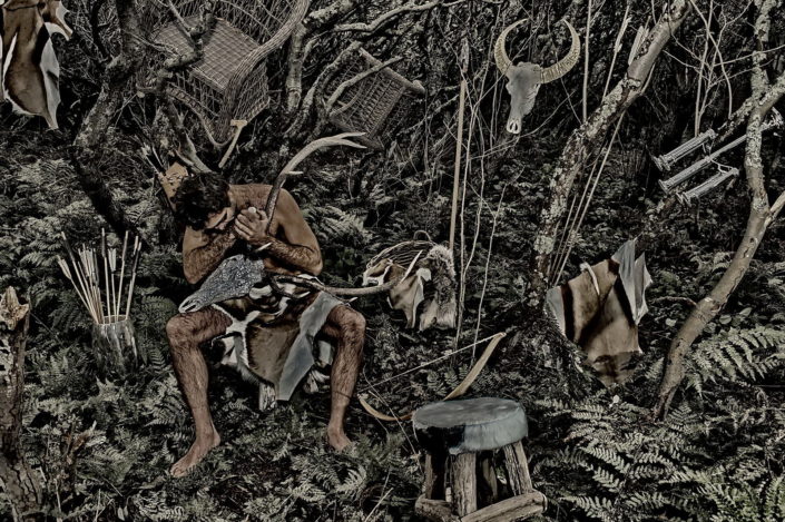 Ein nachkter Man in sylter Wald, nur mit einem Fell bestückt arbeitet und schnitzt einen Swarowski-Geweih. Drumherum sind diverse Gegenstände verteilt