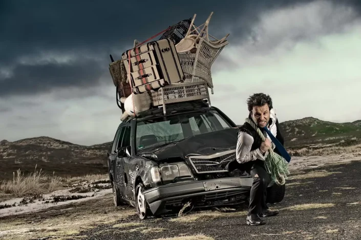 Ein Mann zieht ein mit Möbeln beladenes, beschädigtes Auto durch eine nordische Dünenlandschaft – ein künstlerisches Portrait voller Dramatik.