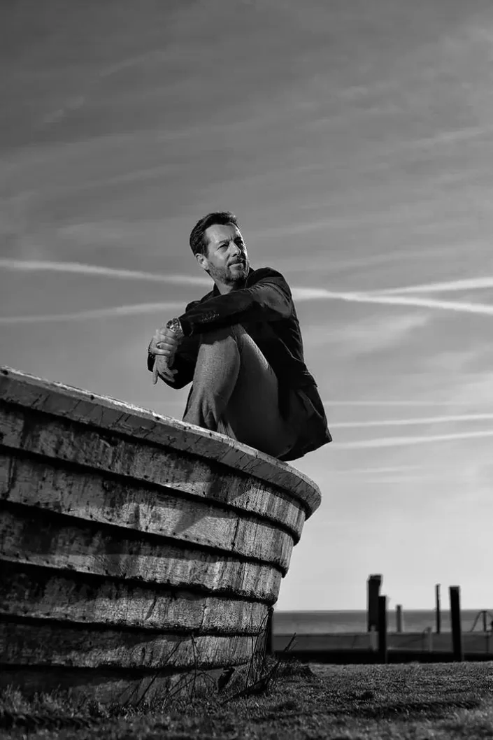 Stilvolles Schwarz-Weiß-Portrait eines Mannes auf einem alten Holzboot an der Küste, Ausdruck von Eleganz und nordischer Gelassenheit.