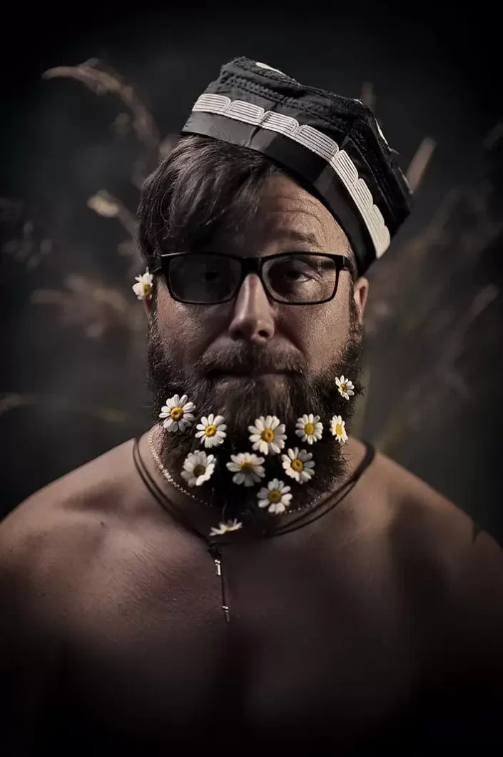 Künstlerisches Portrait eines Mannes mit Brille und Bart, kunstvoll geschmückt mit Gänseblümchen, vor einem eleganten, dunklen Hintergrund.