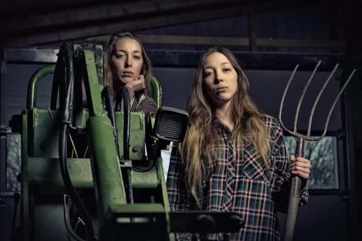 Künstlerisches Portrait zweier Frauen in einer Scheune mit landwirtschaftlichem Gerät und Heugabel, eingefangen in einem eleganten Stil.