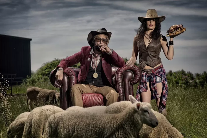 Künstlerisches Portrait im Western-Stil: Ein Paar in Cowboystiefeln und Hüten posiert auf einer Weide mit Schafen für ein exklusives Fotoshooting.