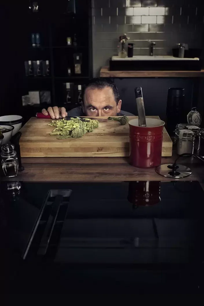 Ein fokussierter Koch blickt hinter einem großen Holzschneidebrett mit Brokkoli hervor – ein stimmungsvolles Portrait für kulinarische Fotografie.
