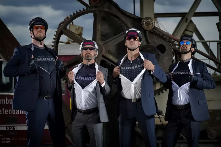 Vier Männer in Anzügen und Radhelmen präsentieren selbstbewusst ihre Triathlon-Trikots bei einem kreativen Fotoshooting auf Sylt.