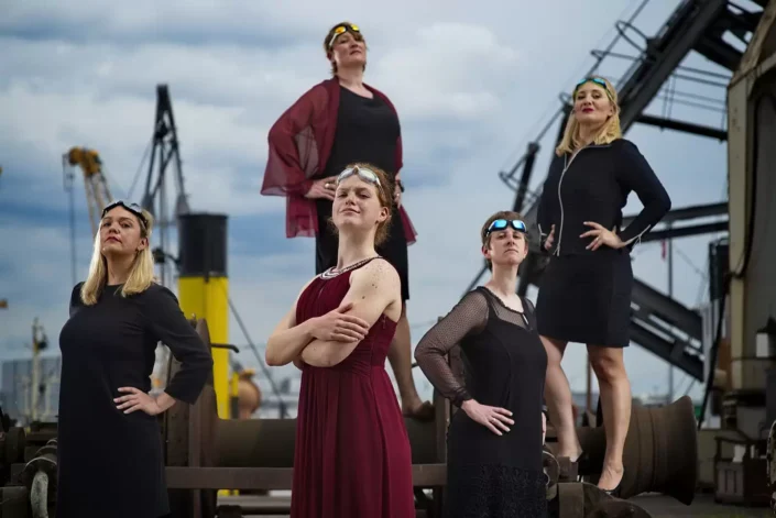 Selbstbewusste Frauen in eleganter Garderobe und mit Schwimmbrillen posieren für ein künstlerisches Portrait vor einer maritimen Hafenkulisse.