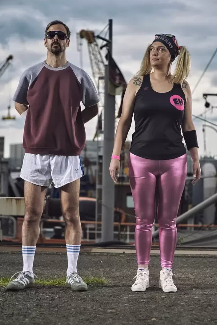 Kreatives Portrait eines Paares im Retro-Sport-Look vor einer Hafen-Kulisse, eingefangen durch professionelle, ausdrucksstarke Fotografie.