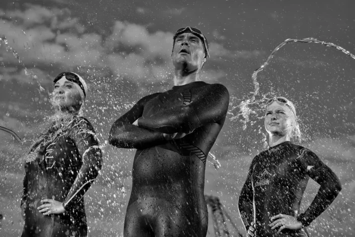 Schwarz-Weiß-Portraits von drei Triathleten in Neoprenanzügen mit spritzendem Wasser, eine kraftvolle Aufnahme für Sportfotografie.