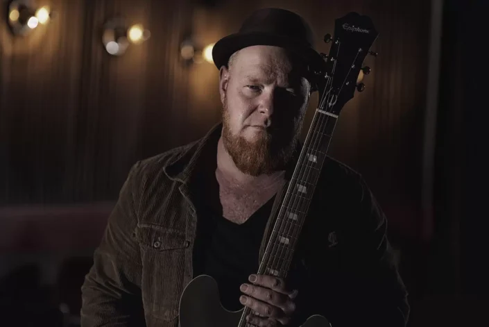 Stimmungsvolles Portrait eines Musikers mit Epiphone-Gitarre und Hut in eleganter, dunkler Lichtsetzung für künstlerische Portraits.