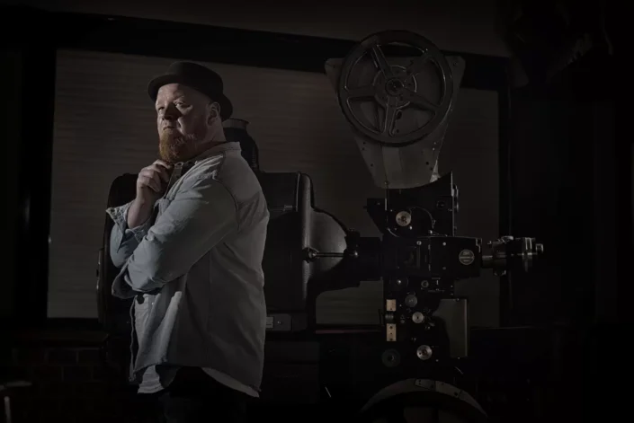 Stilvolles Portrait eines Mannes mit Hut vor einem historischen Filmprojektor, eingefangen in einer atmosphärischen, künstlerischen Lichtsetzung.