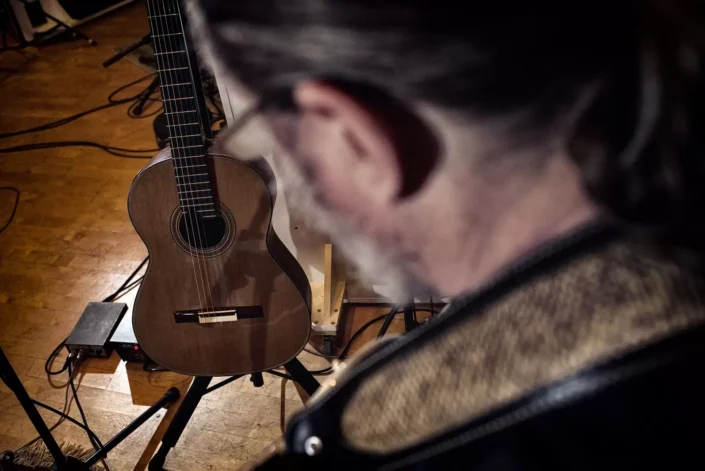 Ein Musiker blickt in einem warm beleuchteten Studio auf eine klassische Konzertgitarre, die auf einem Ständer vor ihm bereitsteht.