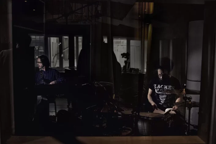 Zwei Musiker in einem abgedunkelten Aufnahmestudio: Ein Mann spielt Klavier, während ein Schlagzeuger konzentriert an seinem Instrument sitzt.