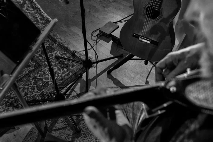 Schwarz-Weiß-Aufnahme einer Gitarre auf einem Ständer, gesehen aus der Perspektive eines Musikers, der ein weiteres Instrument in den Händen hält.