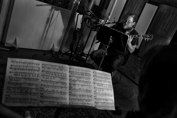 Ein bärtiger Musiker spielt konzentriert Gitarre in einem Studio, während im Vordergrund unscharf ein Notenständer mit Notenblättern zu sehen ist.