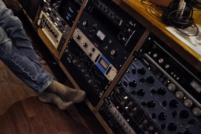 Eine Person in Jeans und Socken sitzt entspannt vor einem Rack mit professionellen Audiogeräten in einem Tonstudio, fotografiert von Fotografen Sylt.
