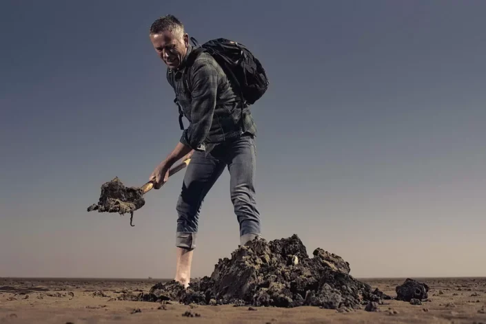Ein Mann mit Rucksack gräbt konzentriert mit einer Schaufel im dunklen Boden einer weiten, kargen Landschaft unter einem klaren, blauen Himmel.