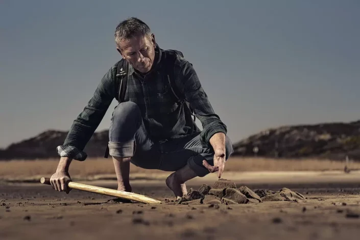 Ein Mann mit Rucksack kniet barfuß im Sand und untersucht konzentriert mit einem Holzstab den Boden in einer ruhigen, natürlichen Umgebung.