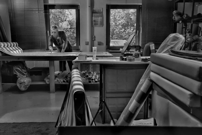 Eine konzentrierte Frau arbeitet in einer Werkstatt an einem großen Tisch, umgeben von Stoffrollen und Materialien in einer ruhigen Atmosphäre.