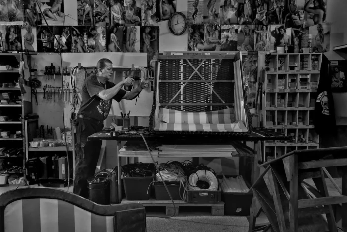 Ein Handwerker arbeitet in seiner Werkstatt konzentriert mit einem Akkuschrauber an einem Polstermöbelstück vor einer Wand voller Fotos.
