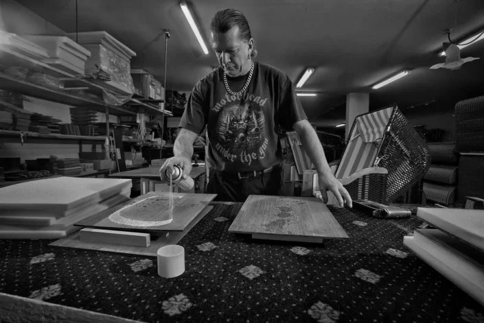 Ein Handwerker in einer Werkstatt sprüht konzentriert Kleber auf ein Holzbrett, während er ein Motorhead T-Shirt trägt. Foto von Fotografen Sylt.