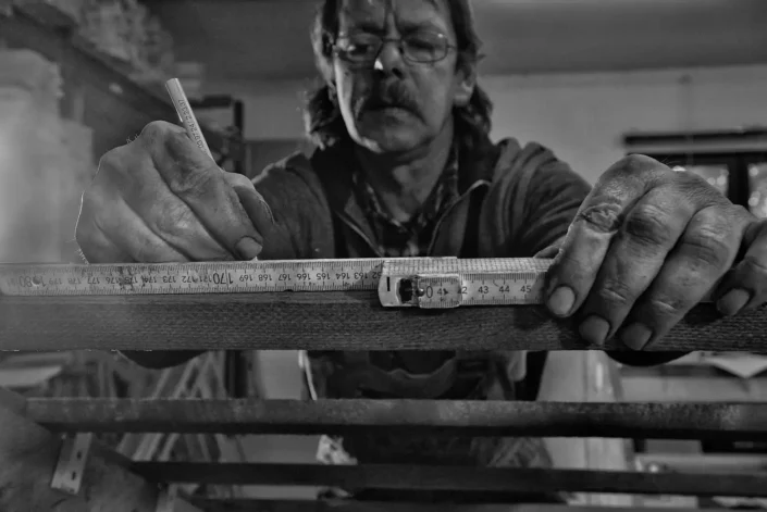 Ein Handwerker mit Brille misst konzentriert ein Holzstück mit einem Zollstock und markiert es sorgfältig mit einem Stift in seiner Werkstatt.