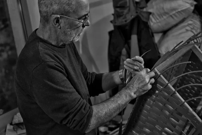 Ein konzentrierter Handwerker mit Brille flicht in einer Schwarz-Weiß-Aufnahme von Fotografen Sylt sorgfältig einen Korb aus schmalen Streifen.