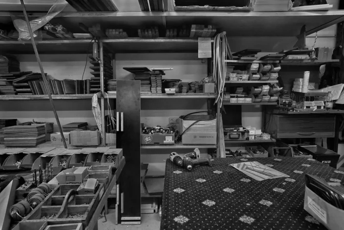 Schwarz-Weiß-Aufnahme einer gut sortierten Werkstatt mit Regalen voller Materialien, Werkzeugen und Kleinteilen auf einem Arbeitstisch.