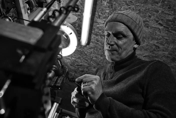 Ein konzentrierter Handwerker mit Mütze arbeitet in seiner Werkstatt an einem feinen Werkstück unter hellem Licht, fotografiert von Fotografen Sylt.