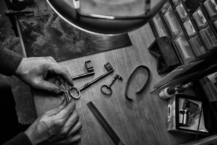 Ein Schlüsseldienst-Handwerker arbeitet in Schwarz-Weiß an einer Werkbank mit alten Schlüsseln und Werkzeugen unter einer hellen Lampe.