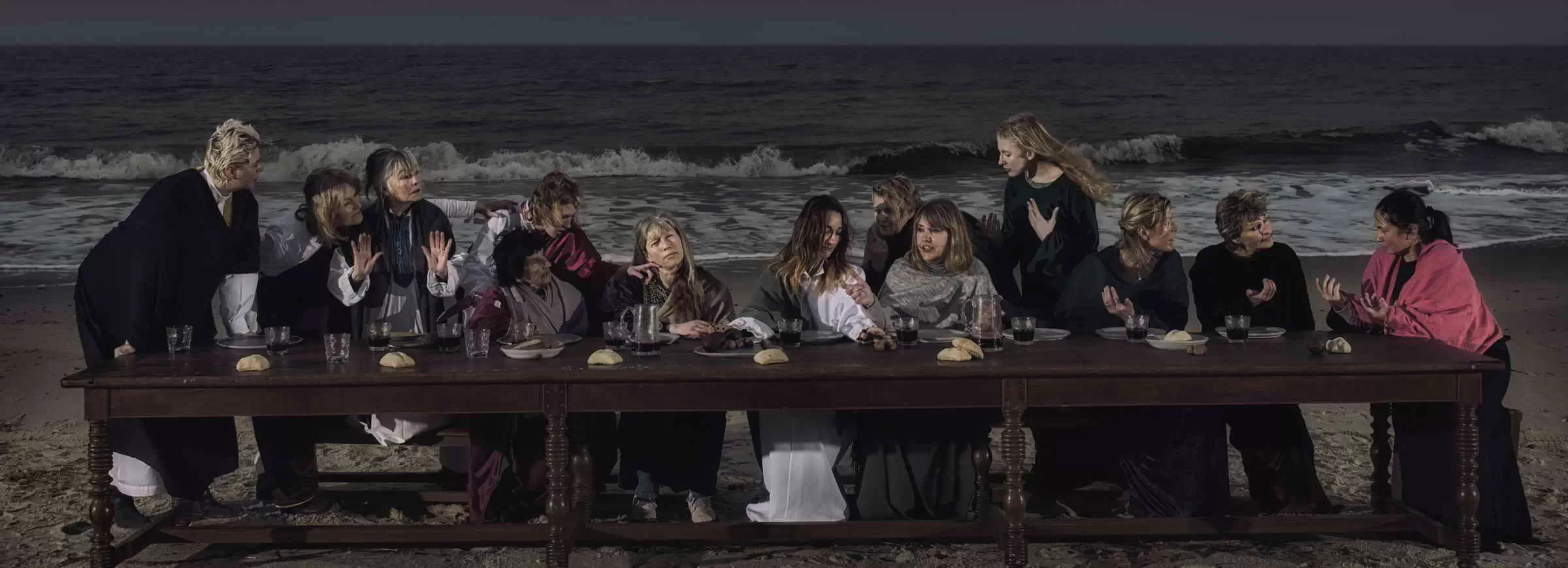 Eine Gruppe von Frauen sitzt an einer langen Tafel am Strand von Sylt in einer Szene, die an das Abendmahl erinnert, bei nächtlicher Stimmung.